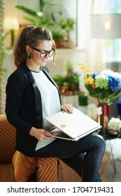 Happy Modern Middle Age Woman In White Blouse And Black Jacket At Modern Home In Sunny Day Sitting On Couch And Opening Laptop.