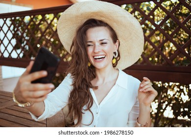 Happy Modern Middle Aged Housewife In White Shirt With Hat Having Web Cam Meeting On A Smartphone In The Terrace.