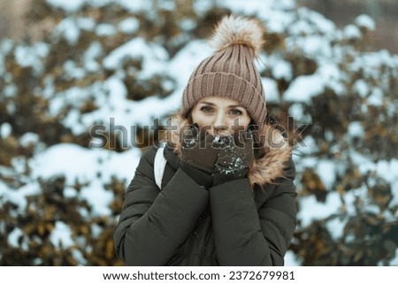 happy modern middle aged woman in green coat and brown hat outdoors in the city park in winter with mittens and beanie hat warming cold hands with breath near snowy branches.