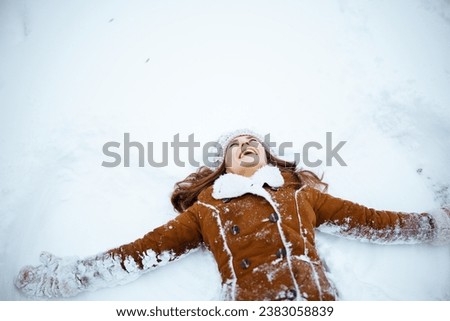 happy modern middle aged woman in brown hat and scarf in sheepskin coat with mittens making snow angels outside in the city in winter.