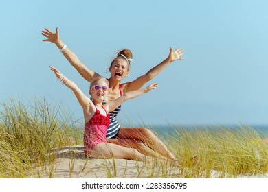 Happy Modern Mother And Child In Beachwear On The Beach Rejoicing. Getting Vitamin D After Long Winter Months. Modern сaucasian Mother 30 Something Years Old In White Blue Striped Swimsuit Brunette.