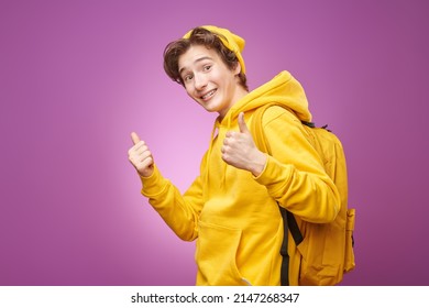 A Happy Modern Teenage Boy In Bright Youth Clothes With Backpack Joyfully Smiles And Shows His Thumbs Up. Purple Background. Youth Fashion And Lifestyle. Bright Youth Colours. Education. 