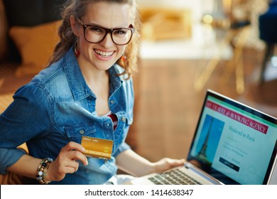 Happy Modern Woman With Opened On Laptop Online Travel & Booking Agency Site Holding Gold Credit Card In The Modern House In Sunny Summer Day. A Fake Website Is Made For Illustrative Purposes.