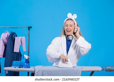 Happy Modern Woman In Shalwar Stands By Laundry Rack Ironing Clothes On Ironing Board Listening To Music In Headphones On Blue Background.