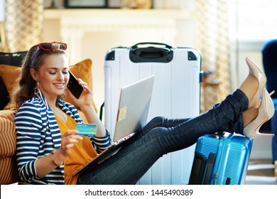 Happy modern woman in striped jacket with credit card buying bus tickets online on a laptop and using ai call center while sitting near couch and trolley bags in the modern living room.