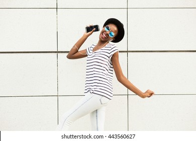 Happy Modern Young Black Woman With Sunglasses, Hat And Striped Shirt  Listening Music Outside And Dancing