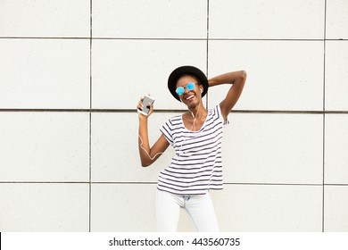 happy modern young black woman with sunglasses, hat and striped shirt  listening music outside and dancing