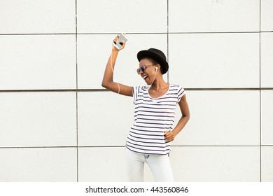 Happy Modern Young Black Woman With Sunglasses, Hat And Striped Shirt  Listening Music Outside And Dancing