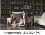 Happy mom and adorable little child girl having fun in play tent, resting on heating floor in handmade toy fort from chairs and blanket, talking, chatting, laughing