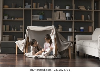 Happy mom and adorable little child girl having fun in play tent, resting on heating floor in handmade toy fort from chairs and blanket, talking, chatting, laughing - Powered by Shutterstock