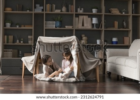 Happy mom and adorable little child girl having fun in play tent, resting on heating floor in handmade toy fort from chairs and blanket, talking, chatting, laughing