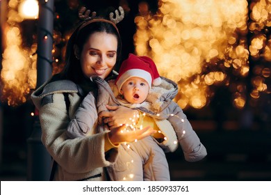 Happy Mom And Baby Holding Christmas Lights Celebrating Outdoors. Happy Mother And Newborn Child Enjoying Winter Holidays 
