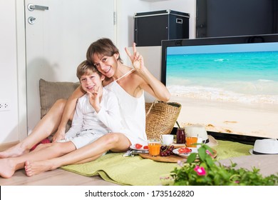 Happy mom and boy imitating summer picnic time on green grass blanket near TV screen. Travelling with family. Coronavirus situation in tourism industry. Quarantine. Stay at home. Isolation.