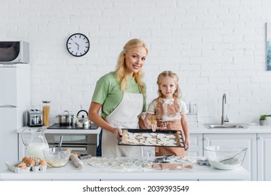 happy mom and child with raw cookies on baking sheet looking at camera in kitchen