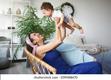 Happy Mom And Child Spending Great Time Together At Home. Attractive Young Hispanic Woman Wearing Blue Dress And Headband Throwing Her Little Son Up, Making Him Laugh. Infancy And Childcare