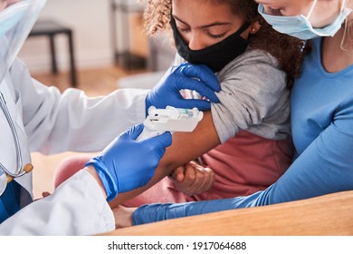 Happy Mom, Child And Young Family Doctor In Medical Face Masks Sitting At The Home. Skilfull Doctor Pointing Modern Electronic Gun Thermometer At Girl's Hand To Measure Her Body Temperature. Epidemic