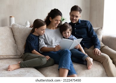 Happy Mom And Dad Telling Fairytale To Cute Sibling Kids. Young Caring Couple Of Parents And Little Children Relaxing On Sofa At Home, Reading Paper Book Together, Enjoying Family Leisure Time