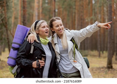 Happy Mom And Daughter Cuddling In The Woods In The Middle Of Shot. Delighted Elderly Woman Holding Trecking Equipment. Young Woman Showing The Way. They Talking.