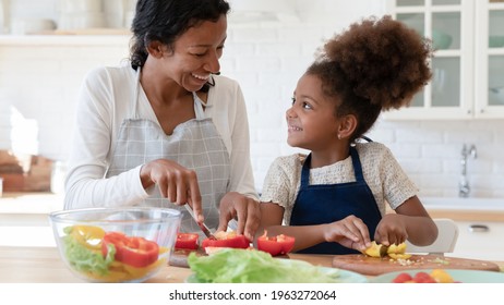 Happy mom and daughter kid talking and laughing while cooking dinner in kitchen together. Sweet preschooler girl helping mom to prepare salad from fresh vegetables, cutting organic pepper on table - Shutterstock ID 1963272064
