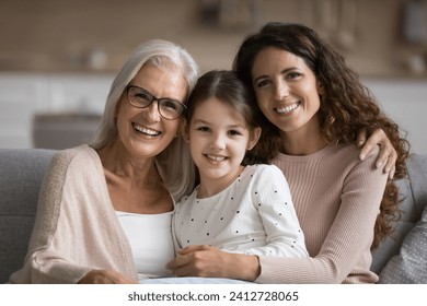 Happy mom and elderly grandma hugging cute tween girl, posing for family portrait on home couch, looking at camera with toothy smiles, enjoying family bonding, leisure together - Powered by Shutterstock