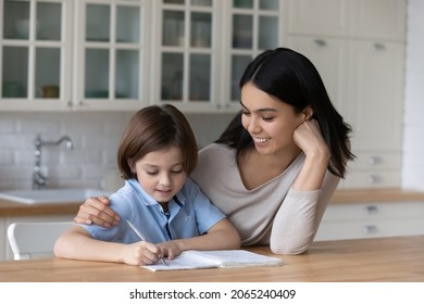 Happy mom giving help   support to schoolboy son while kid doing homework  learning writing exercise  training handwriting in home kitchen  Loving mother encouraging an hugging studying child