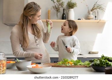 Happy mom giving lettuce leaf to cheerful funny daughter kid for biting and taste. Mother and girl cooking salad in kitchen together, chopping fresh vegetables, organic ingredients, having fun