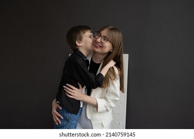 Happy Mom With Glasses And A Preschool-age Son, Hugging And Smiling On A Dark Background. A Family Of Two. Portrait Of Interesting People.