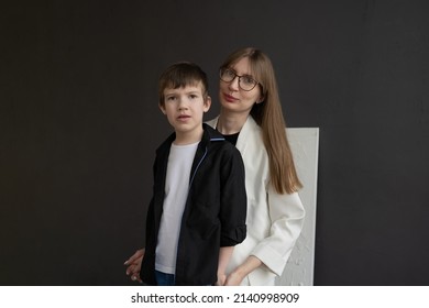 Happy Mom With Glasses And A Preschool-age Son, Hugging And Smiling On A Dark Background. A Family Of Two. Portrait Of Interesting People.