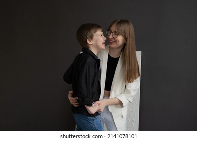 Happy Mom With Glasses And A Preschool-age Son, Hugging And Smiling On A Dark Background. A Family Of Two. Portrait Of Interesting People.