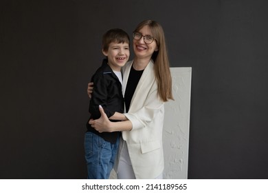 Happy Mom With Glasses And A Preschool-age Son, Hugging And Smiling On A Dark Background. A Family Of Two. Portrait Of Interesting People.