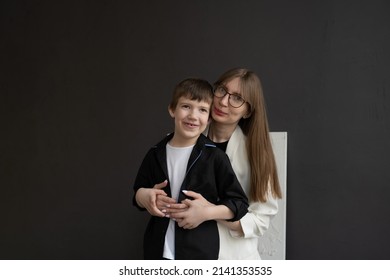 Happy Mom With Glasses And A Preschool-age Son, Hugging And Smiling On A Dark Background. A Family Of Two. Portrait Of Interesting People.