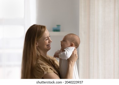 Happy Mom Holding Adorable Baby In Arms, Looking At Little Son, Daughter, Smiling, Laughing. Young Mother Cuddling, Hugging New Born Kid At Home, Enjoying Motherhood. Family, New Parent, Childbirth