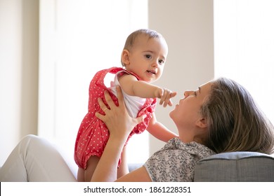 Happy Mom Holding Baby In Arms. Kid Standing On Mothers Lap And Pointing Finger Away. Side View. Parenthood And Childhood Concept