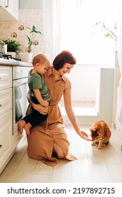 Happy Mom Holding Baby Child Toddler On The Kitchen And Looking On The Domestic Pet Red Cat. Cozy Home And Family Indoors