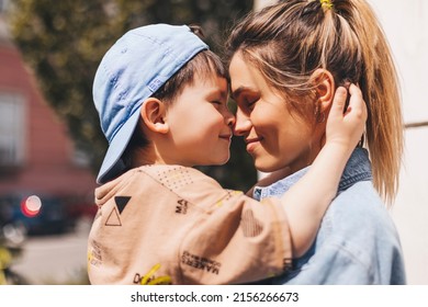 Happy Mom Holding Son Boy In Arms, Hugging Kid With Noses Touch, Smiling, Enjoying Warm Moment. Younger Mum Carrying Kid And Dancing On The Street. Motherhood, Adoption, Family Concept.