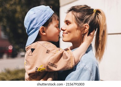 Happy Mom Holding Son Boy In Arms, Hugging Kid With Noses Touch, Smiling, Enjoying Warm Moment. Younger Mum Carrying Kid And Dancing On The Street. Motherhood, Adoption, Family Concept.