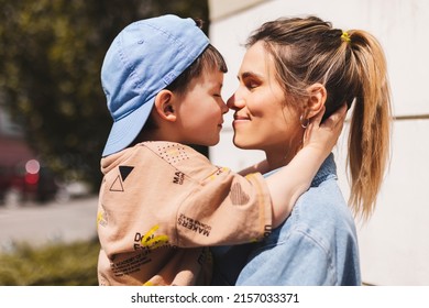 Happy Mom Holding Son Boy In Arms, Hugging Kid With Noses Touch, Smiling, Enjoying Warm Moment. Younger Mum Carrying Kid And Dancing On The Street. Motherhood, Adoption, Family Concept.