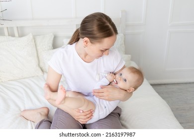 Happy Mom Holds Baby In Her Arms With A Rodent In Her Mouth On A White Bed With Cotton Bedding At Home