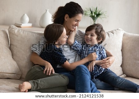 Happy mom hugging little son and daughter with love, care, tenderness, relaxing with kids in couch, talking to children, laughing. Mother and two siblings enjoying leisure at home. Motherhood concept