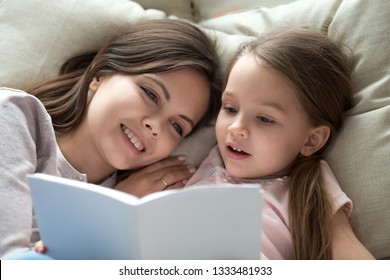 Happy Mom Listening To Little Child Daughter Holding Book Learning Reading Lying In Bed On Pillow, Smiling Mother And Cute Preschool Kid Girl Enjoy Fairy Tale Bedtime Story Together Relax In Bedroom