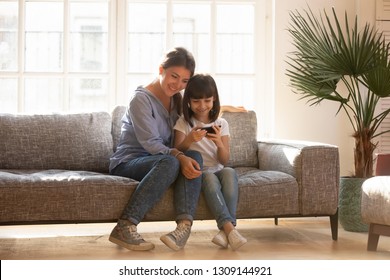 Happy Mom And Little Kid Daughter Enjoy Using Mobile Apps On Smartphone Sitting On Couch, Smiling Mother And Cute Child Girl Play On Phone Gadget Have Fun Together With Cellphone At Home On Sunny Day