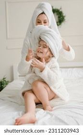 Happy mom puts a towel on girl kid head. Young mother with cute little daughter in bathrobe, towels on heads sitting lovely on bed. Healthcare and family concept. Cheerful beauty day at home. Closeup. - Shutterstock ID 2311203943