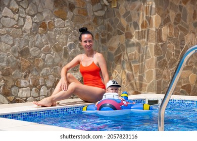 Happy Mom In A Red Swimsuit With Her Little Son Are Playing Outdoors, Against The Backdrop Of A Pool With Blue Water.