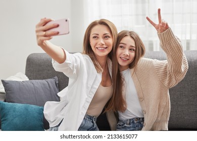 Happy Mom Sitting Close To Smiling Teen Daughter Showing Two Fingers Gesture While Taking Self Portrait On Smartphone On Couch At Home