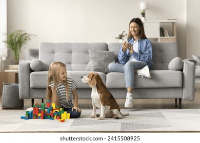 Happy mom taking picture of pet and child, sitting on comfortable sofa, watching little kid and dog playing with building cubes on warm floor, using mobile phone, shooting, recording - Powered by Shutterstock