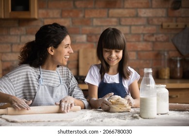 Happy mom teaching cute daughter girl to bake, rolling, kneading dough, talking to kid, smiling, laughing. Funny child with flour on face helping mom to cook pastry for family dinner