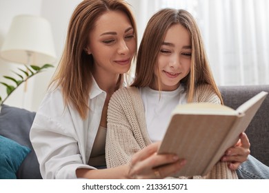 Happy Mom And Teen Daughter Smiling And Reading Interesting Book While Relaxing On Couch On Weekend Day At Home