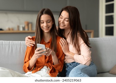 Happy mom with teen daughter using phone together at home, watching social media videos, doing online shopping on smartphone sitting on sofa together - Powered by Shutterstock