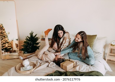 A Happy Mom With Two Daughters Is Resting, Relaxing And Having Fun Sitting On The Bed In A Cozy Decorated Bedroom During The Christmas Holiday At Home During The New Year Vacations. Selective Focus