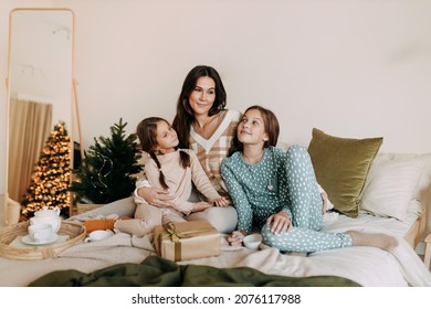 A Happy Mom With Two Daughters Is Resting, Relaxing And Having Fun Sitting On The Bed In A Cozy Decorated Bedroom During The Christmas Holiday At Home During The New Year Vacations. Selective Focus